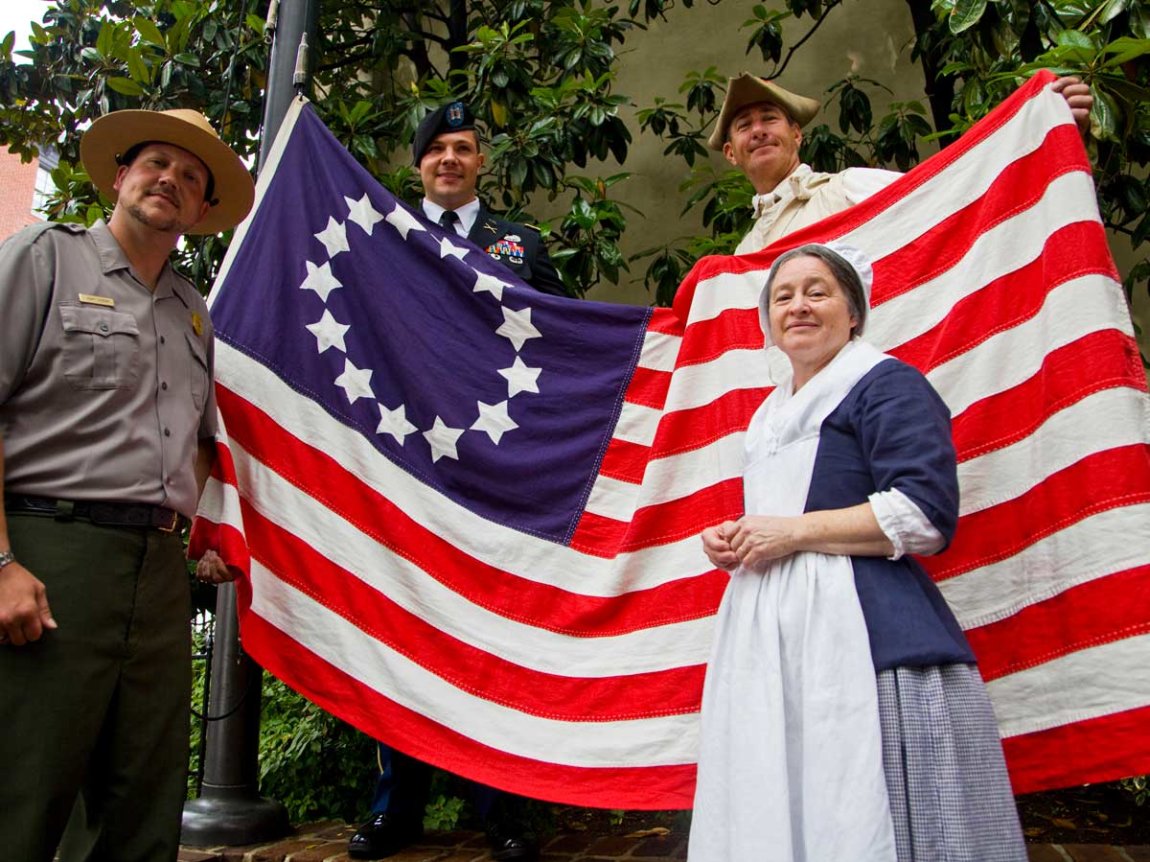 four people in colonial dress unfurl an early-design American flag